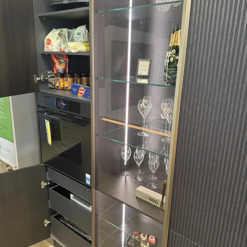 Nolte NEO Ausstellungsküche: geöffneter Vorratshochschrank neben beleuchteter Glasvitrine und Einbaubackofen