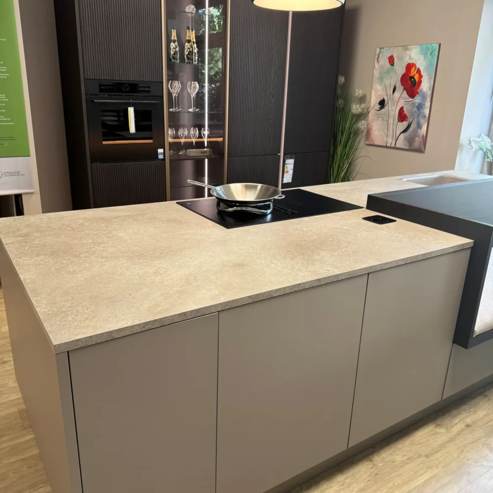 Nolte NEO Ausstellungsküche: Kochinsel mit Lackfronten Lava matt und Arbeitsplatte Sandstein Creme, dahinter Hochschrankwand mit Glasvitrine